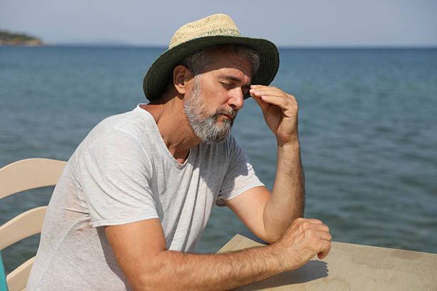 Middle Aged Man Wearing Straw Hat Sitting By The Sea With Eyes Closed And Hands On Temples, Expressing Stress, Headache, Fatigue, Or Mental Overload During Summer Vacation, Highlighting Tension, Burnout, And Emotional Pressure In Leisure Environment.