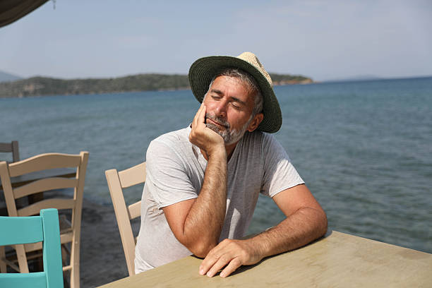Middle Aged Man Wearing Straw Hat Sitting By The Sea With Eyes Closed And Head Resting On Hand, Peaceful Summer Vacation Scene, Relaxed Mood, Calm Emotion, Leisure Time And Mindful Moment Near Ocean.