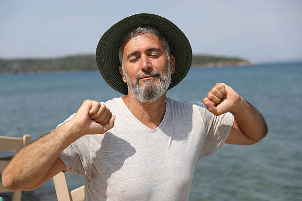 Middle Aged Man Wearing Straw Hat Standing By Sea With Eyes Closed And Raised Fists, Expression Of Relaxation And Satisfaction, Summer Vacation Moment, Calm Mood, Freedom And Positive Emotion By Ocean.