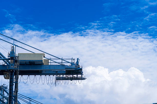Large blue and white crane is suspended in the air, with a clear blue sky in the background. The crane is positioned high above the ground, and its blue