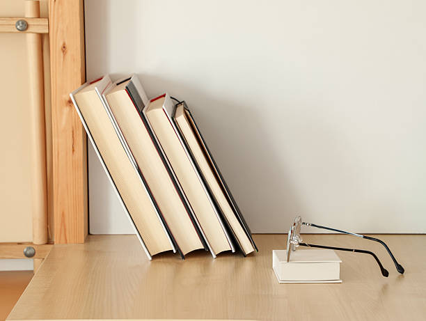 Reading glasses placed on a bright wooden table next to a row of books. Ideal for studying, educational activities, reading sessions, or creating a warm, inviting home environment.