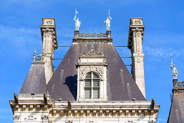 Steep slate roof with ornate stonework and classical statues holding spears on the upper facade of Hotel de Ville in Paris, set against a vivid blue sky and highlighted by crisp daylight.