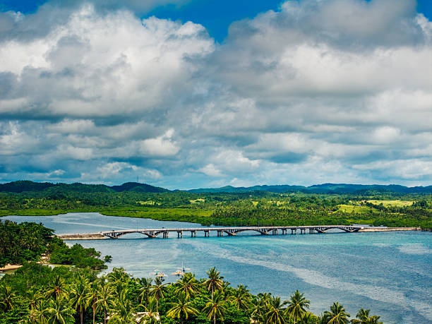 A vivid tropical landscape on Siargao Island, Philippines featuring a long bridge across calm waters, lush palm-lined shores, and rolling green hills under a dramatic cloudy sky.