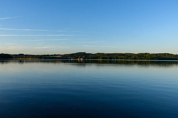 Glasslike surface of Lac des Settons reflects the distant tree-lined shore and soft evening light, with tranquil blue tones and gentle ripples creating a peaceful atmosphere.