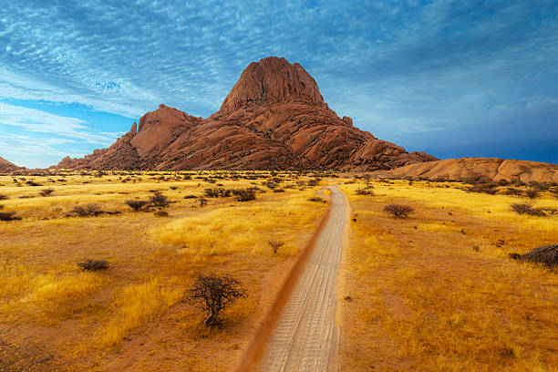 A dirt road winds through golden grass toward the towering Spitzkoppe in Namibia, with a dramatic red rock peak against a wide blue sky. A remote, adventurous landscape inviting exploration and travel.