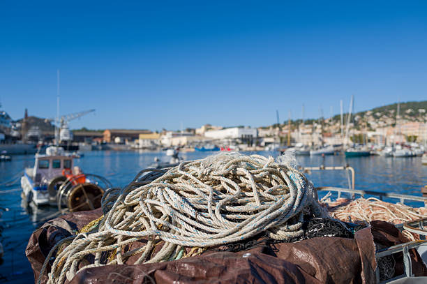 Close-up of coiled fishing ropes and nets resting on a moored boat in La Ciotat harbor, with a sunlit marina, blue sky, and blurred waterfront buildings in the background. The scene highlights maritime textures and a relaxed coastal atmosphere.