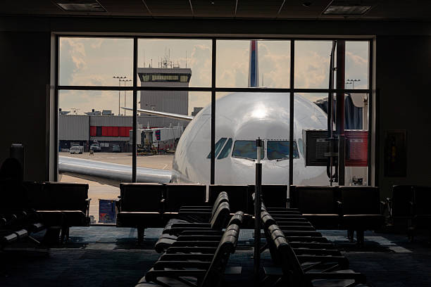 View of a commercial airplane parked at an airport gate, seen through large terminal windows with empty waiting seats in the foreground, conveying travel, transportation, and anticipation before departure.