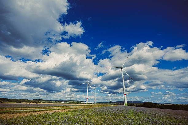 Wind turbines rotate in a field under a dramatic sky filled with fluffy clouds. The scene captures the beauty of renewable energy in a peaceful rural setting.