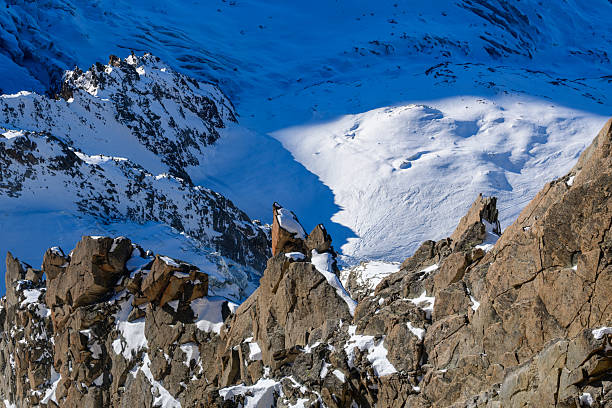 Jagged brown rock formations rise above a sweeping glacier blanketed in snow, with deep blue shadows creating dramatic contrast in the Mont Blanc massif.