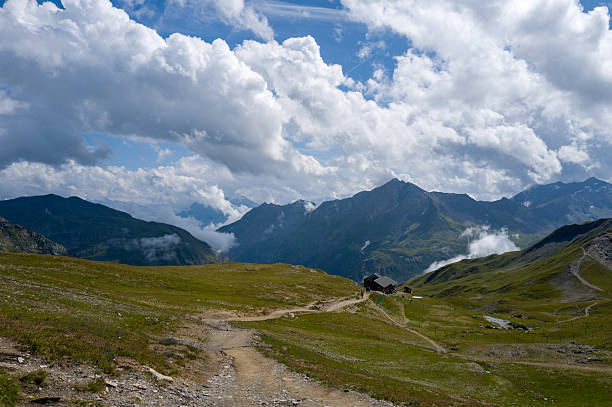 A dirt hiking trail leads to a small mountain refuge on a grassy plateau, surrounded by dramatic peaks and dynamic clouds in the French Alps.
