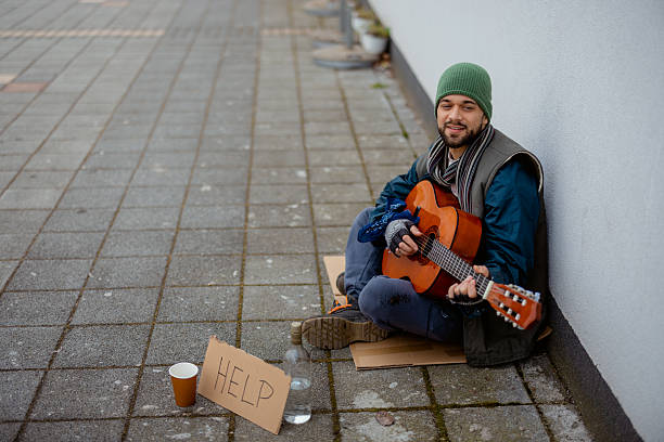 Homeless adult man sitting on a city street during cold weather while playing guitar. Represents homelessness, poverty, and social issues in urban environments.