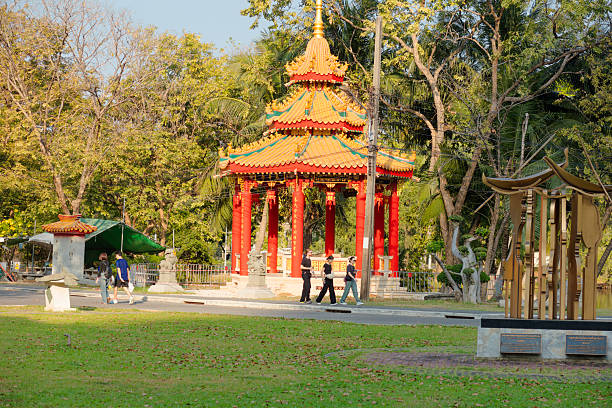White young tourists are passing by a red chinese pavilion in Lumpini Park in Bangkok downtown at afternoon. Around are many tropcal trees. View from one pathway to other pathway through park. There are women and a man with red beard. Two of them are wearing sunglasses. In left area a thai couple is walking.
