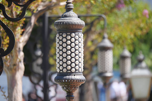 A close-up photo of a small, ornate Moroccan lantern on a red background. The lantern is made of metal with colorful glass panels and intricate details. It is a popular decorative item used during Ramadan and other Islamic festivals
