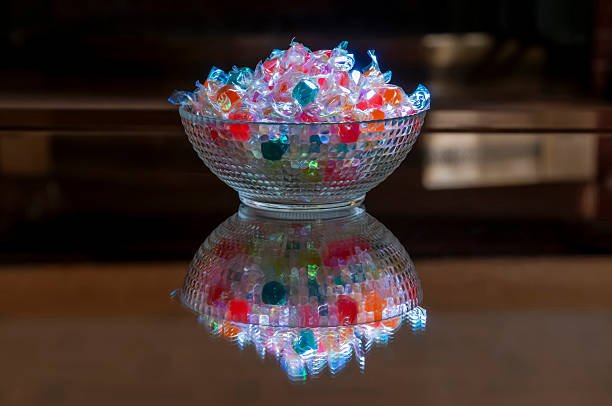 A glass bowl filled with vibrant, colorful candies and sweets that emit a soft glow, perfectly reflected on a dark table in a celebratory setting.