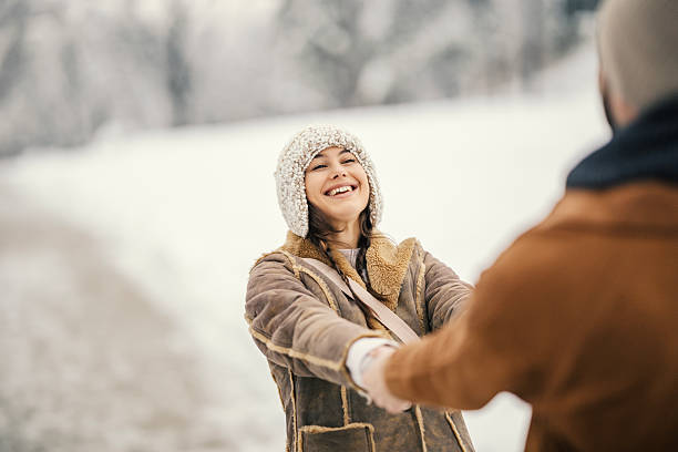 Young woman wearing a warm sheepskin coat and knit hat, smiling while holding hands with her partner against a snowy winter background, expressing joy and connection