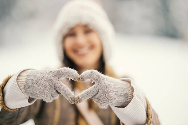 Young woman wearing warm winter clothing and smiling with a blurred background, making a heart shape with her gloved hands, symbolizing love and care in cold weather