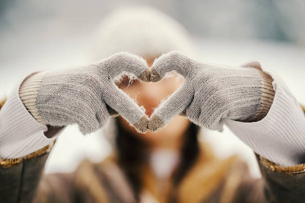 Person wearing warm gloves forming a heart shape with their hands, expressing love and coziness during cold winter weather, with a soft bokeh background