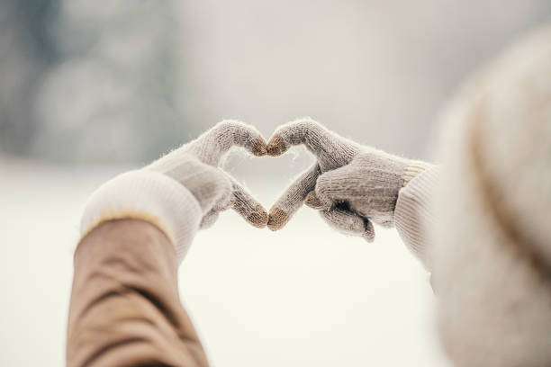 Person wearing winter gloves forming a heart shape with fingers against a soft snowy background, representing love, warmth, and connection in cold weather