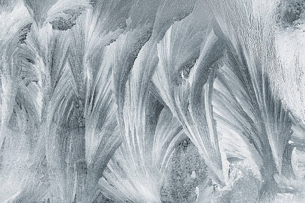 A close-up view reveals intricate frost patterns spreading across a window pane, creating a delicate and beautiful display of nature's artistry during the winter season. The crystalline texture of the ice forms feathery shapes, perfect for backgrounds or seasonal themes.