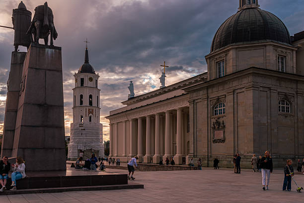 Vilnius, Lithuania – August 23, 2025: Cathedral Square at sunset with the Gediminas monument, Vilnius Cathedral, and Bell Tower visible. People gather in the open space under dramatic evening clouds.