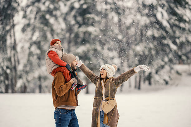 Happy family with a young boy on his father's shoulders and mother with arms open, playing together outside in a snowy winter forest, feeling joy and bonding