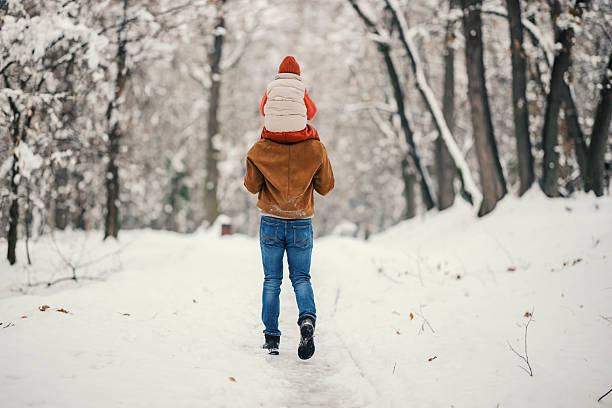 Father gives son a piggyback ride along a snow-covered forest path, sharing a playful, protective moment of bonding and outdoor winter adventure in warm clothing