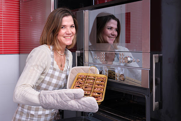 Smiling woman wearing apron and oven mitts pulling a tray of brownie bites from a kitchen oven