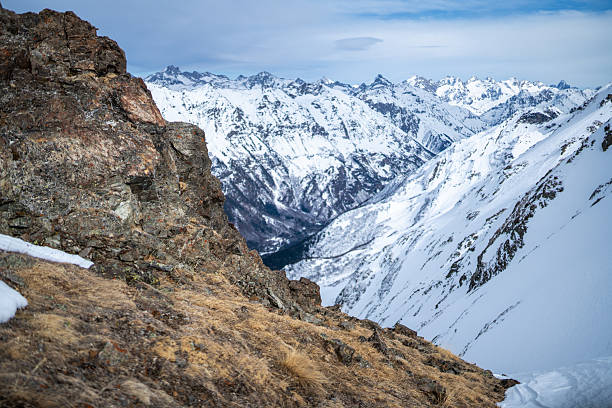 Snow-covered alpine mountains rise behind a rocky foreground, capturing a vast, tranquil wilderness. A dramatic, cold landscape perfect for travel, nature, and outdoor adventure themes.