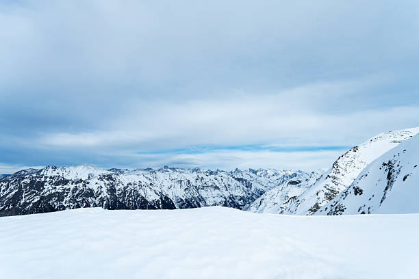 A tranquil alpine landscape featuring a vast snowfield and rugged mountain silhouettes under a pale blue sky. Ideal for travel, nature, winter sports, and outdoor adventure themes.