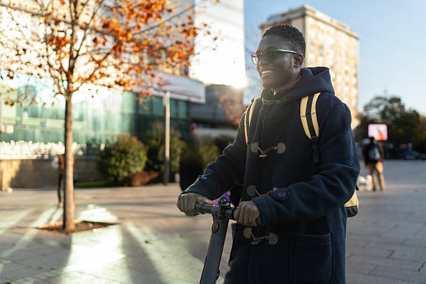 Young black man in sunglasses and a duffel coat smiles while riding an electric scooter on a sunny urban street in autumn, backpacked and enjoying micro-mobility freedom