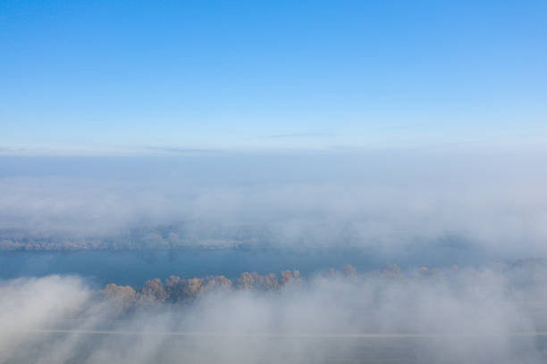 Aerial view of a tranquil river partially obscured by drifting fog, with bare winter trees and a clear blue sky above. The soft light and ethereal atmosphere evoke calm and serenity.
