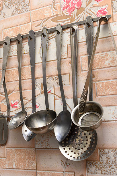 row of steel kitchen utensils including ladles of various sizes and skimmers suspended on a metal bar against a wall with a floral ornament in the interior