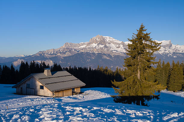 Panoramic winter scene of snow covered La Clusaz village nestled in a hilly landscape in Annecy, France. Wooden cabins are scattered across the snowy hillside, surrounded by snow laden trees