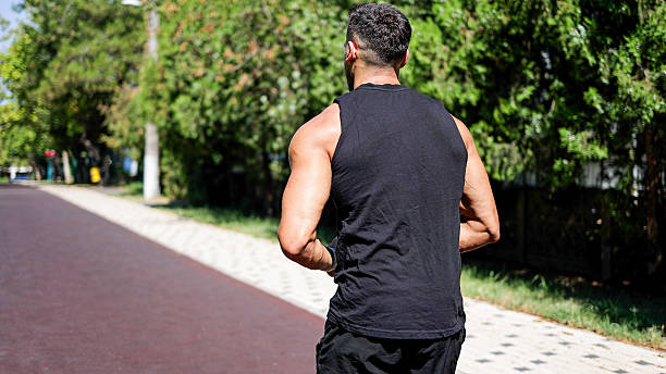 The image shows a man running along a paved path in a park area, with trees and greenery lining both sides of the walkway. Sunlight enhances the vivid colors of the surroundings, while the man's athletic build and focused expression highlight themes of endurance, outdoor exercise, and personal fitness goals. Elements of the park such as benches, vegetation, and open space support concepts related to healthy routines, nature-based workouts, and urban recreational environments.