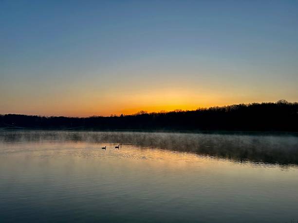 Sunrise and a clear sky reflect off a lake in the early morning. As light breaks over the trees and water a light fog sets in over the warming landscape.