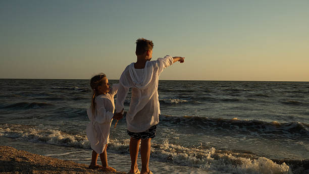 Two siblings stand together on a sandy beach at sunset, holding hands and pointing toward the open ocean. The children share a moment of admiration and connection with nature on the seashore..
