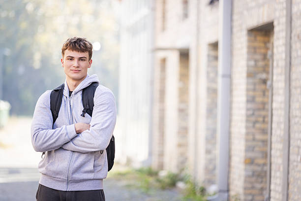 A young man in a gray hoodie with a backpack stands outdoors, arms crossed, by a brick building. The bright, casual campus vibe conveys readiness, confidence, and everyday student life.