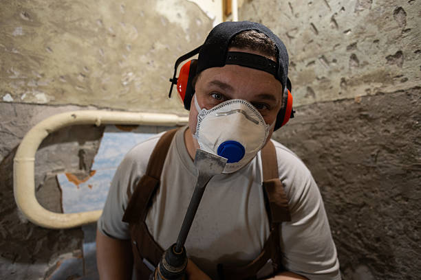 Man worker wearing a respirator mask, hearing protection, and cap, holding a drill tool, looking at the camera while removing old plaster from a wall during renovation work..