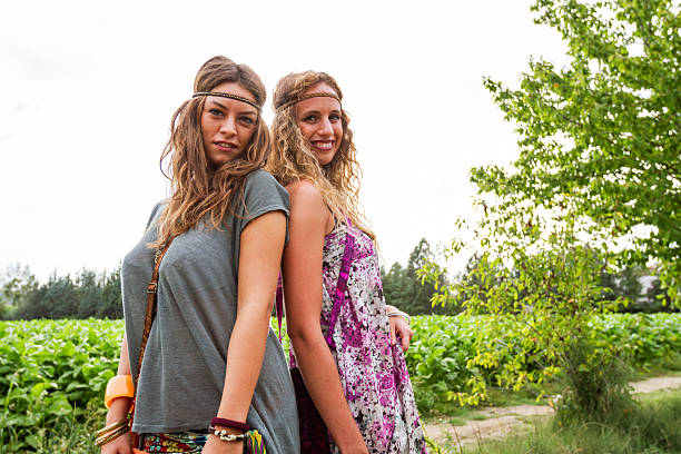 Two young women standing back to back outdoors in a green natural landscape, wearing casual bohemian clothing and headbands, expressing friendship, confidence, and relaxed lifestyle in a summer setting.