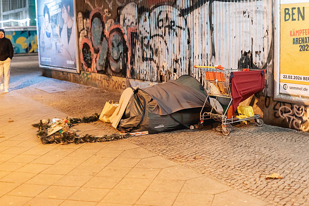 Berlin, Germany – January 1, 2026: A makeshift shelter with a covered sleeping area, a trolley, and personal belongings stands beside a graffiti wall with posters at night.
