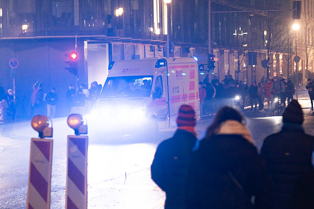 Berlin, Germany – January 1, 2026: An ASB ambulance with flashing lights approaches a street intersection as a crowd watches on a wet roadway at night.