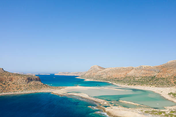 Expansive sandy lagoon with vivid turquoise and deep blue waters framed by rugged hills at Balos Beach, Crete, Greece. The scene is sunlit and open, with clear skies and natural textures.