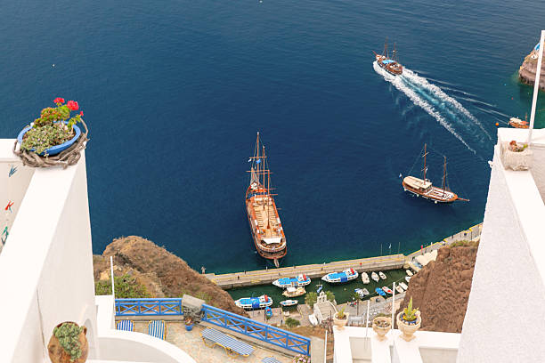 Traditional sailing boats and yachts at the harbor below white architecture in Santorini, Greece, seen from above.