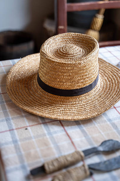 A high-angle, close-up shot of a vintage-style straw hat with a black ribbon band, resting on a rustic checkered tablecloth. In the background, blurred primitive hand tools and a wooden interior create an authentic, historical, or rural atmosphere. Perfect for themes of traditional craftsmanship, summer fashion, gardening, or historical reenactment.