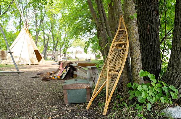 A pair of authentic, hand-crafted wooden snowshoes with rawhide webbing leaning against a mossy tree trunk. The background features a sprawling green summer field, a distant historical white building, and a bright sky. This image captures the intersection of traditional Indigenous craftsmanship and North American frontier history, evoking themes of winter survival, outdoor adventure, and heritage tools in a serene natural landscape