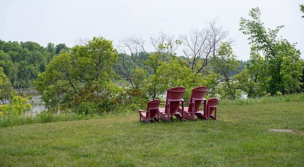 A peaceful summer scene featuring a family set of four red wooden Adirondack (Muskoka) chairs—two adult-sized and two child-sized—arranged on a lush green lawn. The chairs face a scenic river or lake bordered by dense green trees under a soft, overcast sky. This high-angle shot evokes feelings of relaxation, family vacation, and the quiet beauty of a riverside retreat, making it ideal for travel, lifestyle, and real estate marketing.