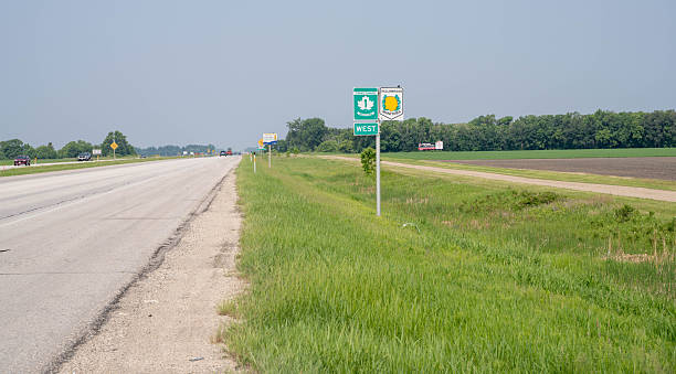 Railroad crossing sign stands prominently in rural Lamar, Colorado. Massive grain silo dominates the scene, surrounded by industrial buildings and structures. Power lines and transmission towers stretch across flat, grassy landscape with dirt roads. Bright blue sky with scattered clouds completes quintessential small-town American vista.