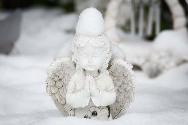 Close-up of a white cherub angel figurine praying in a snowy garden. A pile of snow sits like a hat on its head. Peaceful winter scene.