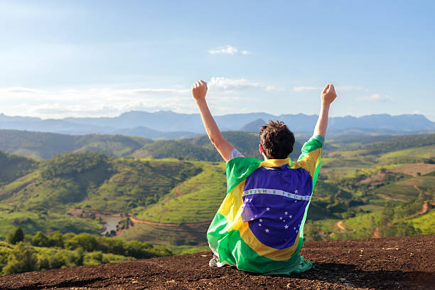 Brazilian fan sitting on a mountain with raised arms and national flag, expressing national pride and passionate supporters spirit. Concept of football support, unity, strength and love for Brazil.