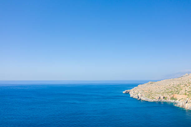 Vast expanse of deep blue Mediterranean water meeting a sunlit, arid rocky coastline on the southern edge of Crete. The scene is tranquil and open, with clear skies and minimal vegetation along the rugged shore.